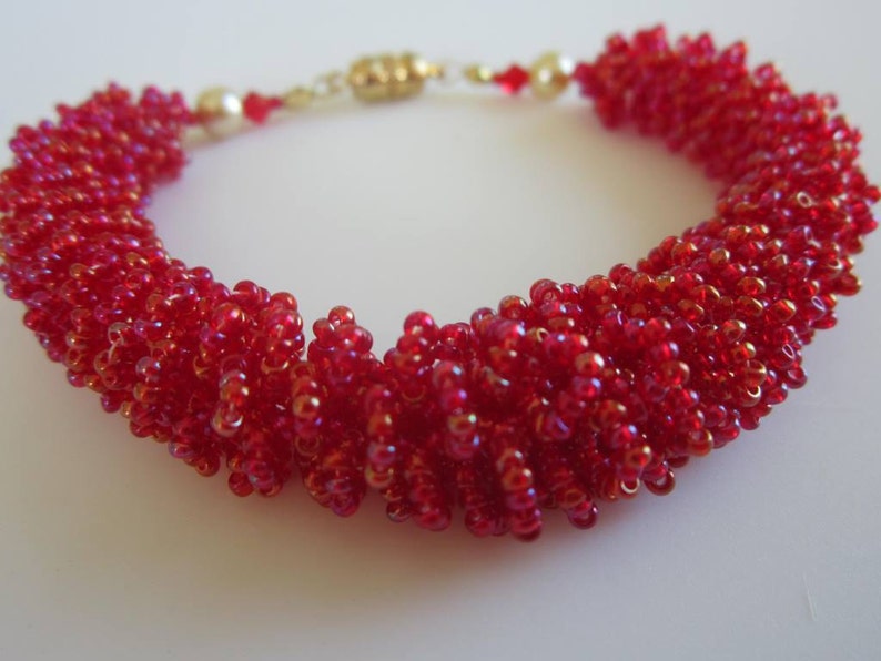May include: A red beaded bracelet with a gold clasp. The beads are small and iridescent, creating a shimmering effect.