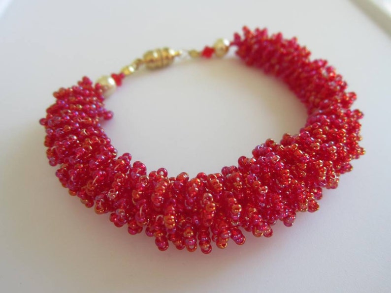 May include: A red beaded bracelet with a gold clasp. The beads are small and tightly packed, creating a textured look.