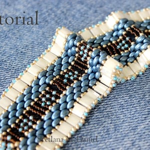 May include: A close-up of a beaded bracelet with a blue, white, and brown color scheme. The bracelet is made with small, round beads and larger, rectangular beads. The bracelet is lying on a blue denim fabric.