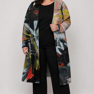May include: A long-sleeved open-front duster coat featuring an abstract expressionist print with bold strokes of black, white, yellow, and red. The garment is worn over a solid black scoop-neck top and matching black wide-leg trousers with black boots.