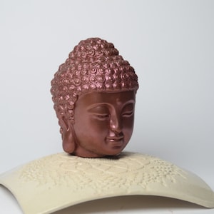 May include: A copper-colored Buddha head sculpture with closed eyes and a serene expression. The head is placed on a white, decorative base.