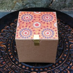 May include: A square gift box with a brown cardboard exterior and a colorful patterned top. The pattern features red, orange, blue, and white circular designs.