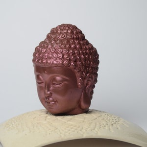 May include: A copper-colored Buddha head sculpture with closed eyes, sitting on a white, decorative base.