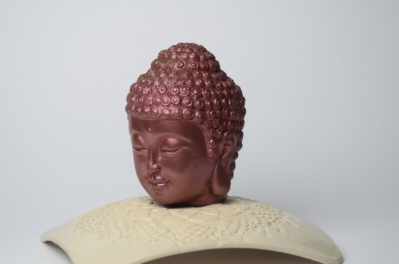 May include: A copper-colored Buddha head sculpture with closed eyes, sitting on a white, decorative base.