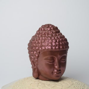 May include: A copper-colored Buddha head sculpture with a detailed, textured hairstyle. The head is facing forward with closed eyes and a serene expression.