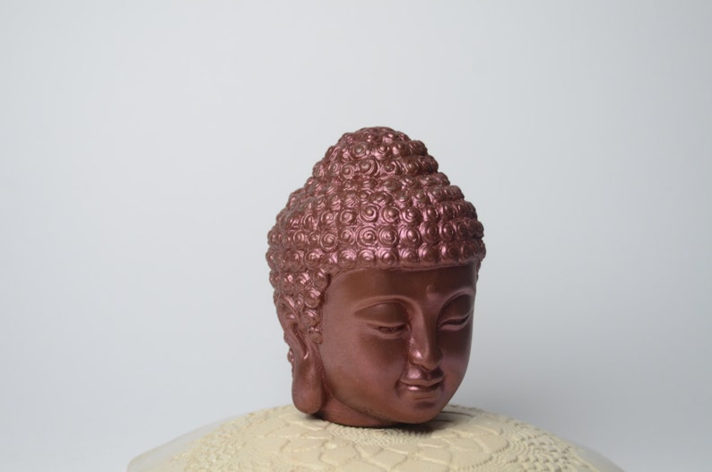 May include: A copper-colored Buddha head sculpture with a detailed, textured hairstyle. The head is facing forward with closed eyes and a serene expression.