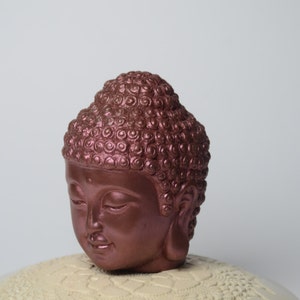 May include: A copper-colored Buddha head statue with a detailed, textured hairstyle. The statue is sitting on a white, decorative base.