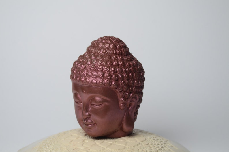 May include: A copper-colored Buddha head statue with a detailed, textured hairstyle. The statue is sitting on a white, decorative base.