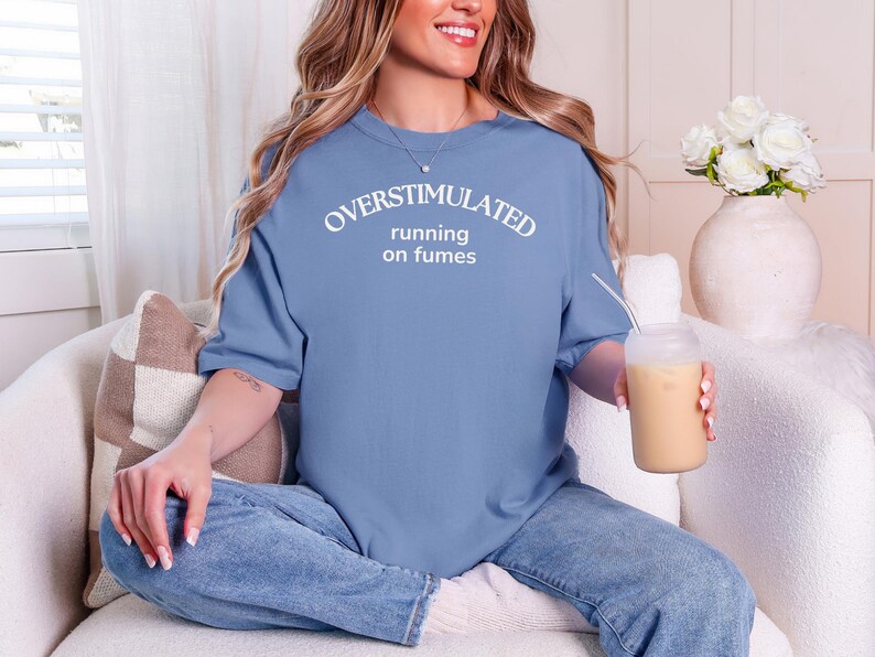 May include: A pale blue t-shirt with the text "OVERSTIMULATED running on fumes" in white. The shirt is worn with blue jeans. A clear glass with a straw and a beige drink is held.