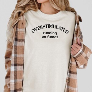 May include: A cream-coloured t-shirt with the text "OVERSTIMULATED running on fumes" in black. The person is wearing a brown and tan plaid flannel shirt over the t-shirt.