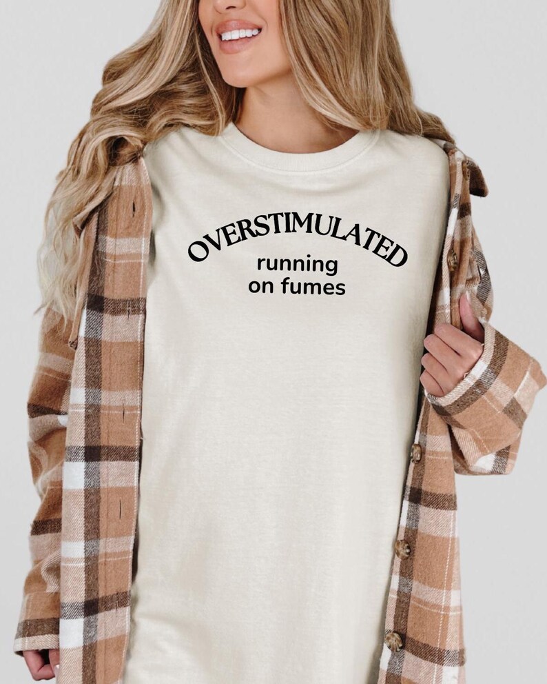May include: A cream-coloured t-shirt with the text "OVERSTIMULATED running on fumes" in black. The person is wearing a brown and tan plaid flannel shirt over the t-shirt.