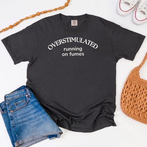 May include: A charcoal grey t-shirt with the white text "OVERSTIMULATED running on fumes". The image includes blue denim shorts, white trainers, and a woven brown handbag. A wooden beaded necklace is also present.