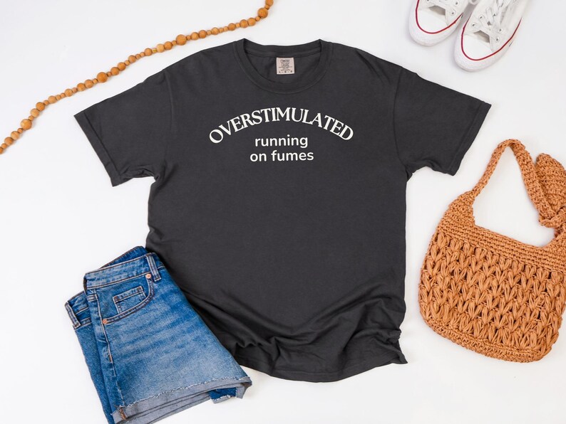 May include: A charcoal grey t-shirt with the white text "OVERSTIMULATED running on fumes". The image includes blue denim shorts, white trainers, and a woven brown handbag. A wooden beaded necklace is also present.