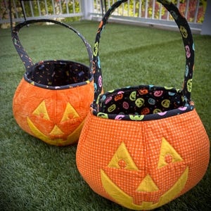 Peut inclure: Deux paniers en tissu orange en forme de citrouille avec des traits de visage jaunes. Les paniers ont une doublure noire avec des visages de jack-o'-lantern colorés et des poignées assorties. Les paniers sont sur de l'herbe verte.