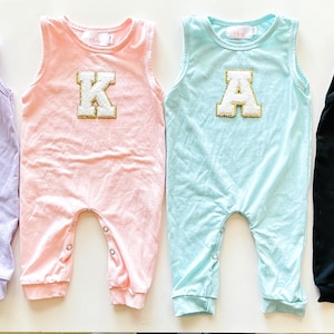 Peut inclure: Quatre barboteuses pour bébés aux couleurs pastel : lavande avec un cœur, rose avec un "K", bleu clair avec un "A" et noir avec un "L". Chacune a un design sans manches et des fermetures à boutons-pression en bas.