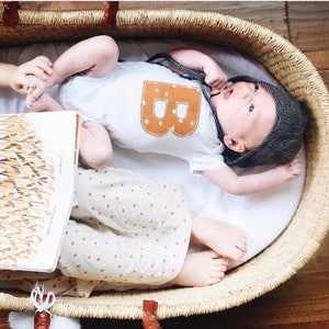 May include: A baby wearing a white onesie with a brown felt letter B and stars on the front, lying in a woven basket with a book and another child's legs.