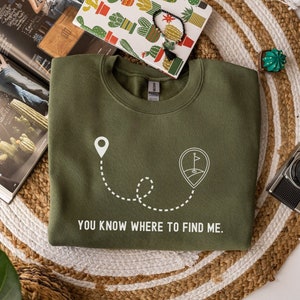 May include: Olive green sweatshirt with a white graphic of a location pin, a dotted line, and a golf course pin. The text "YOU KNOW WHERE TO FIND ME" is printed in white. The sweatshirt is displayed on a woven mat.