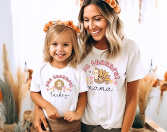 May include: A white t-shirt with a pink and brown turkey graphic and the text "one thankful turkey" on it. A second white t-shirt with a pink and brown turkey graphic and the text "one thankful mama" on it. Both shirts have a floral design around the turkey graphic.