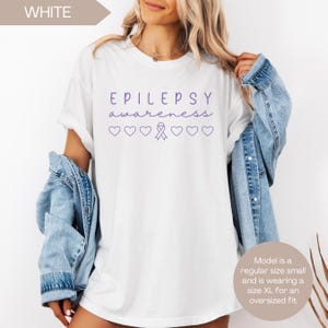 Pu&ograve; includere: T-shirt bianca con le parole "EPILEPSY awareness" in corsivo viola, insieme a cuori e un nastro viola. La maglietta &egrave; una taglia piccola regolare, ma viene indossata come una XL oversize.