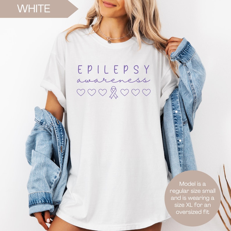 Pu&ograve; includere: T-shirt bianca con le parole "EPILEPSY awareness" in corsivo viola, insieme a cuori e un nastro viola. La maglietta &egrave; una taglia piccola regolare, ma viene indossata come una XL oversize.