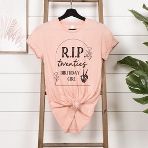 Op de afbeelding: Een perzikkleurig T-shirt met een zwarte afbeelding waarop staat "RIP twenties birthday girl" met een skelet hand die een peace-teken maakt.