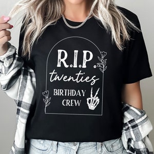 Op de afbeelding: Zwart T-shirt met een witte afbeelding van een grafsteen met de tekst "RIP twenties Birthday Crew" met een skelet hand die het vredesteken geeft.