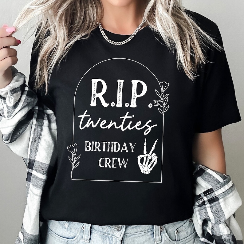 Op de afbeelding: Zwart T-shirt met een witte afbeelding van een grafsteen met de tekst "RIP twenties Birthday Crew" met een skelet hand die het vredesteken geeft.