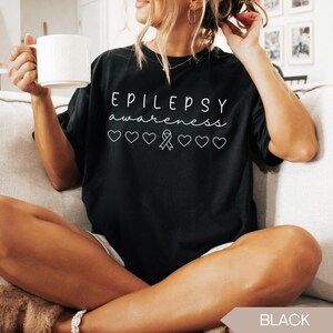 Pu&ograve; includere: T-shirt nera con le parole "EPILEPSY awareness" in corsivo bianco. Sotto il testo ci sono cinque cuori bianchi e un nastro di sensibilizzazione bianco. La maglietta &egrave; in stile casual.