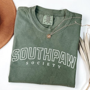 May include: A green t-shirt with the text "Southpaw Society" in white outlined letters. The shirt is made of a soft, comfortable fabric and has a relaxed fit.