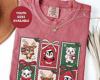 May include: A red Comfort Colors t-shirt with a festive design featuring cartoon animals in postage stamp frames. The design includes a reindeer, penguin, and polar bear, each holding books or wearing Santa hats. The text "YOUTH SIZES AVAILABLE" is in a red circle.