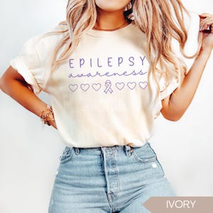 Pu&ograve; includere: T-shirt color avorio con le parole "EPILEPSY awareness" in corsivo viola. Sotto il testo ci sono cuori viola e un nastro di sensibilizzazione viola. La maglietta &egrave; abbinata a jeans blu.