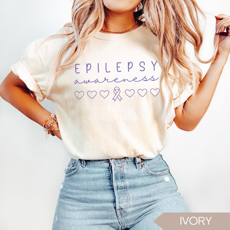 Pu&ograve; includere: T-shirt color avorio con le parole "EPILEPSY awareness" in corsivo viola. Sotto il testo ci sono cuori viola e un nastro di sensibilizzazione viola. La maglietta &egrave; abbinata a jeans blu.