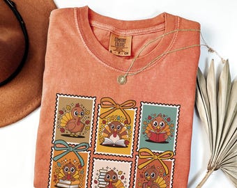 May include: Orange Comfort Colors t-shirt featuring a graphic of six postage stamp-style images of cartoon turkeys. Each turkey is depicted in various poses, some with books and bows. A gold necklace and a brown hat are also visible.