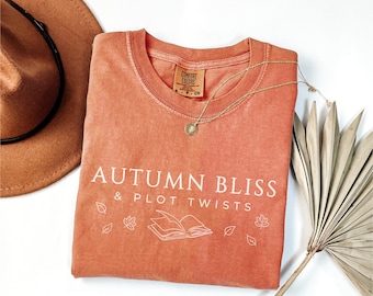 May include: A burnt orange t-shirt with the text "Autumn Bliss & Plot Twists" in white. The shirt features a small illustration of a book and leaves.
