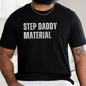 K&ouml;nnte beinhalten: Schwarzes T-Shirt mit wei&szlig;em Text, der "Step Daddy Material" lautet.