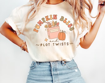 May include: Cream-colored t-shirt with the text "Pumpkin Bliss and Plot Twists" in a retro font. The graphic includes a book, pumpkin, and fall foliage. The shirt is paired with blue jeans.