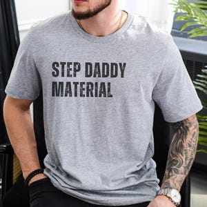 K&ouml;nnte beinhalten: Ein graues T-Shirt mit dem Aufdruck "Step Daddy Material" in Schwarz.