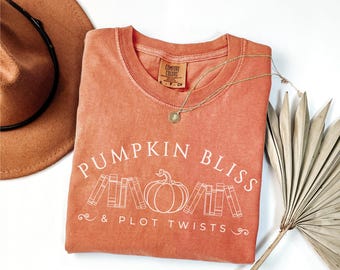 May include: A burnt orange t-shirt with the text "Pumpkin Bliss & Plot Twists" in white. The design features a pumpkin with stacked books on either side.