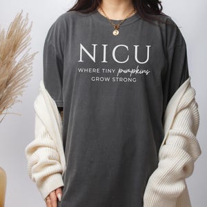 Puede incluir: Una camiseta gris oscuro con el texto "NICU Where Tiny Pumpkins Grow Strong" impreso en blanco.