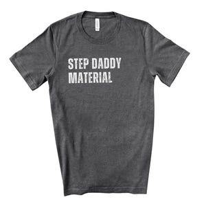 K&ouml;nnte beinhalten: Ein dunkelgraues T-Shirt mit dem Aufdruck "Step Daddy Material" in Wei&szlig;.