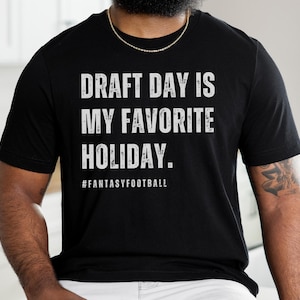 May include: A black t-shirt with white text that reads "Draft Day is My Favorite Holiday."  The text is in a distressed font.  The hashtag #FantasyFootball is printed below the text.