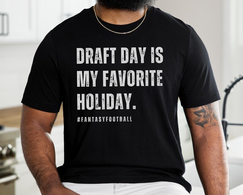 May include: A black t-shirt with white text that reads "Draft Day is My Favorite Holiday."  The text is in a distressed font.  The hashtag #FantasyFootball is printed below the text.