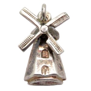 May include: A silver-toned metal charm shaped like a windmill, featuring four rotating blades attached to a central point. The main body of the windmill has a tapered, conical structure with two small, rectangular window-like cutouts on the front.