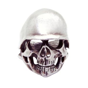 May include: A silver-colored skull ring with detailed facial features, including eye sockets, a nose cavity, and teeth. The ring has a smooth, rounded top and a hollowed-out interior.