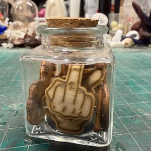 May include: A clear glass jar with a cork stopper, containing wooden cutouts of a hand giving the middle finger. The jar is square and sits on a green surface. The wooden cutouts are light brown.