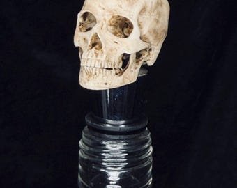 Puede incluir: Un tapón de botella con una calavera humana realista de color blanco hueso con detalles marrones desgastados. La calavera está montada sobre una base cónica de cromo pulido con una junta de goma negra, insertada en el cuello de una botella de vidrio.