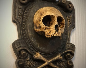 May include: A decorative wall plaque featuring a gold-painted skull with crossed bones, set against a dark brown background. The skull is framed by a gold-painted ornate design with a crown at the top.