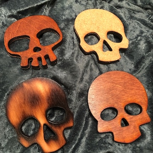 May include: Four wooden skull coasters in different shades of brown. The coasters are shaped like a skull with a simple design.