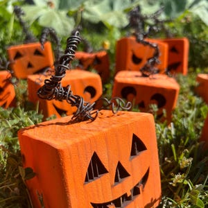 Könnte beinhalten: Orangefarbene Holz-Jack-o'-Lanterns mit geschnitzten Gesichtern und Drahtstielen sitzen im grünen Gras. Die würfelförmigen Kürbisse haben dreieckige Augen und einen lächelnden Mund, ideal für Halloween-Dekorationen.