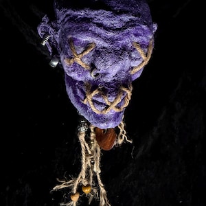 May include: A handcrafted, purple fabric doll head with stitched-on rope details for eyes and mouth. The doll has a textured surface and is adorned with dangling twine, beads, and a wooden pendant. The background is black.